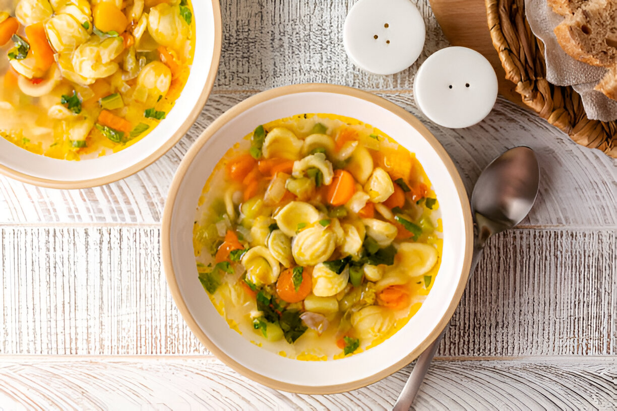 A fresh bowl of vegetable soup with pasta served on a wooden table, accompanied by a spoon and a basket of bread.