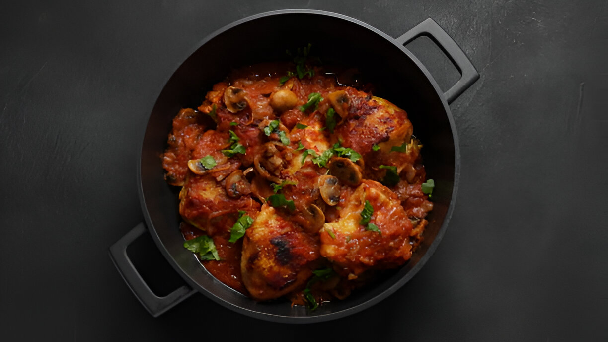 A black pot filled with chicken pieces, vibrant red tomato sauce, and sliced mushrooms, garnished with fresh parsley.