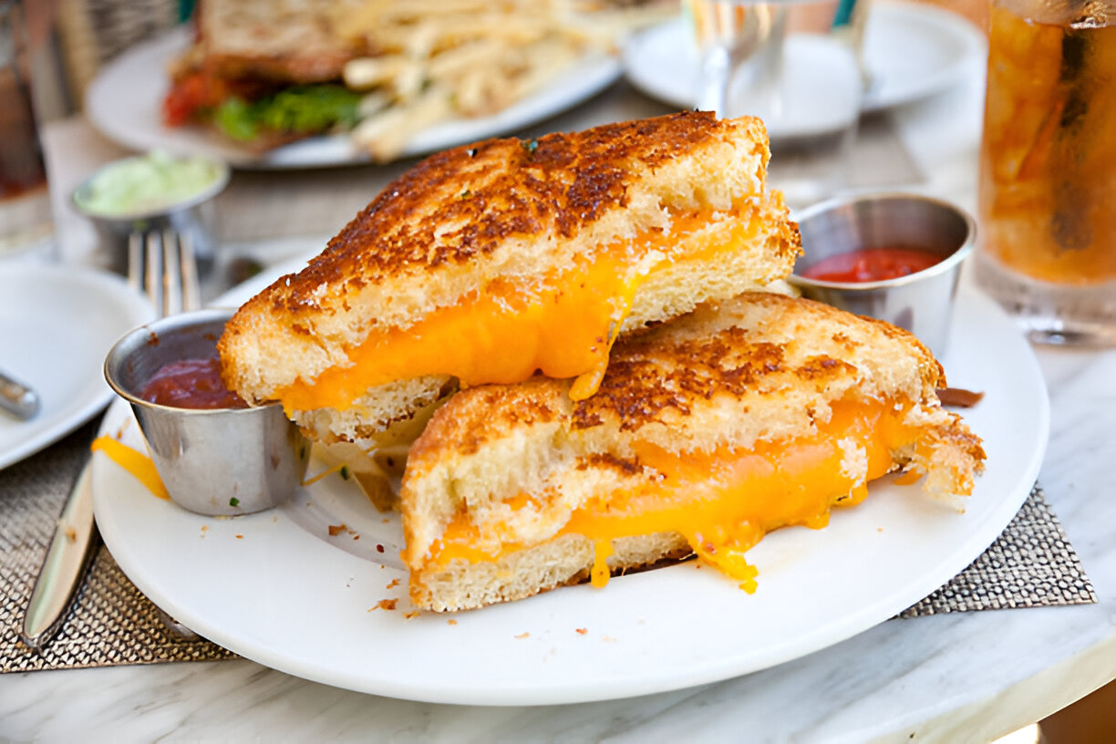 A delicious grilled cheese sandwich with melted cheddar cheese spilling out, served with a side of dipping sauce.