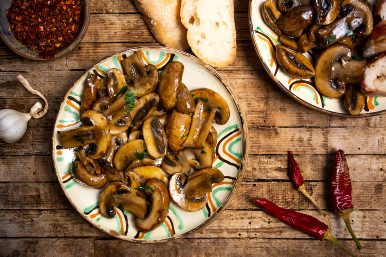 A colorful plate of sautéed mushrooms with garlic and herbs, accompanied by red chili peppers and slices of bread on a rustic wooden surface.