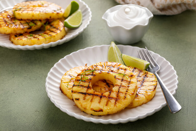 Grilled pineapple slices served with lime wedges and a dollop of cream on a green background.