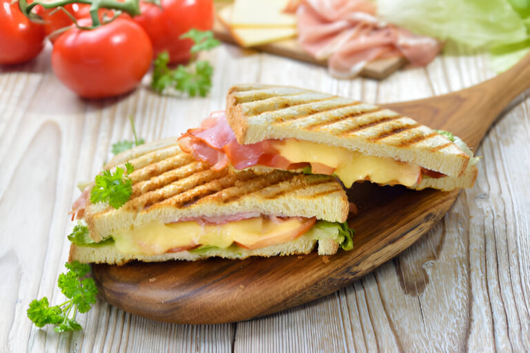 Grilled sandwich with cheese, ham, and lettuce on a wooden board, surrounded by fresh tomatoes and herbs.