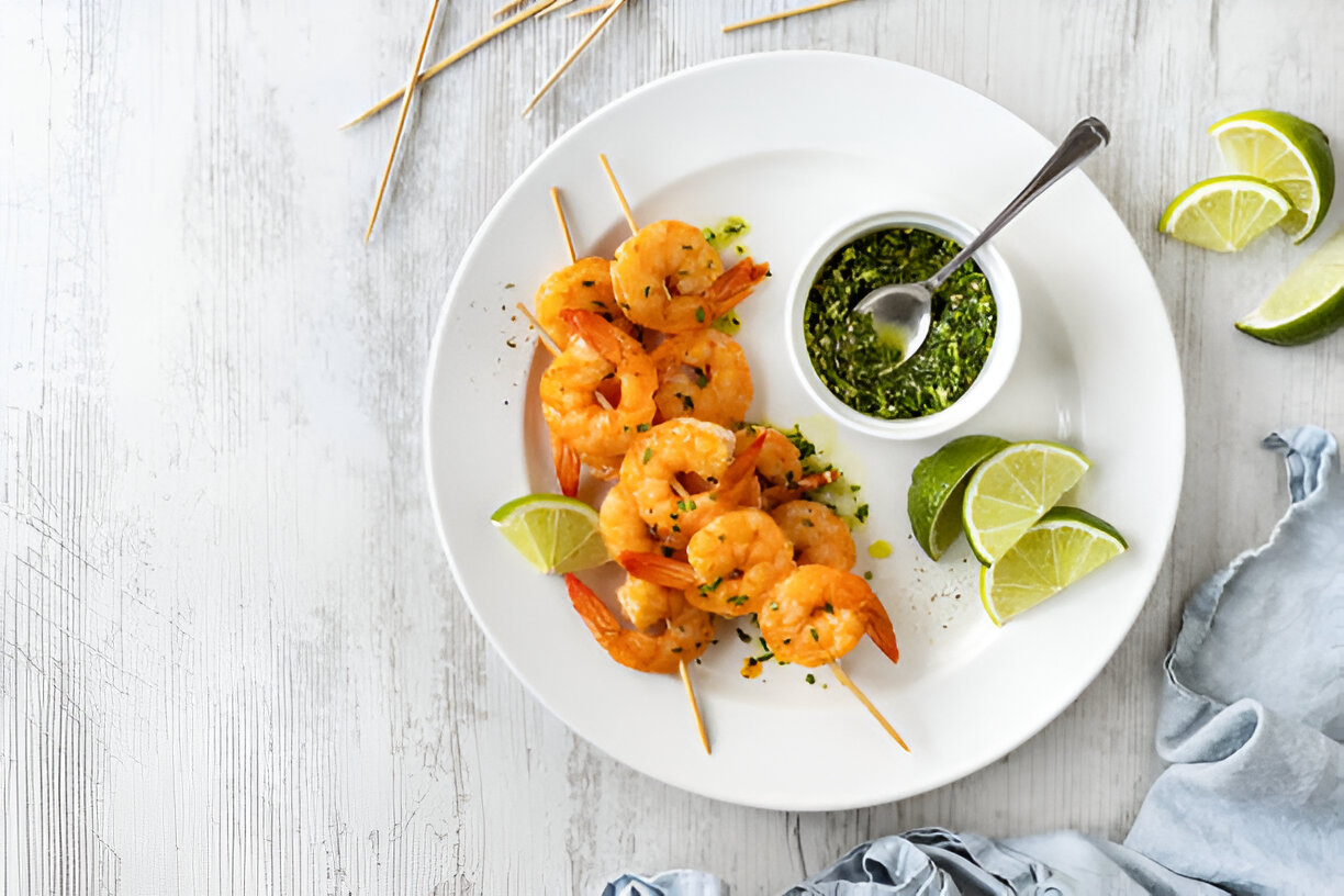 Grilled shrimp skewers served on a white plate with lime wedges and a small bowl of green sauce.
