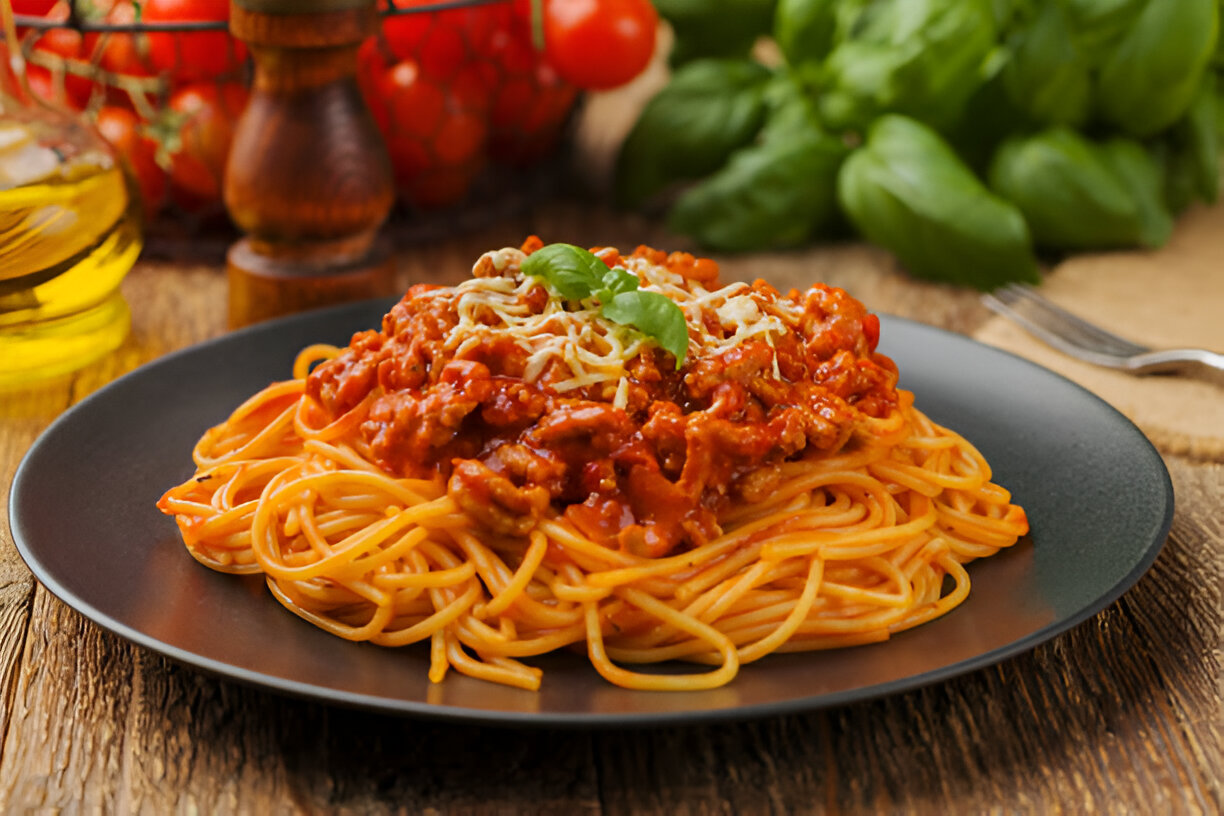 A plate of spaghetti topped with meat sauce, garnished with basil leaves and grated cheese, with tomatoes and basil in the background.