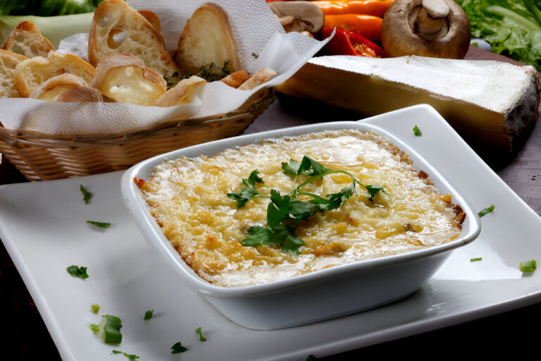 Creamy baked dish with cheese and herbs served in a white bowl, accompanied by a basket of bread and various vegetables.