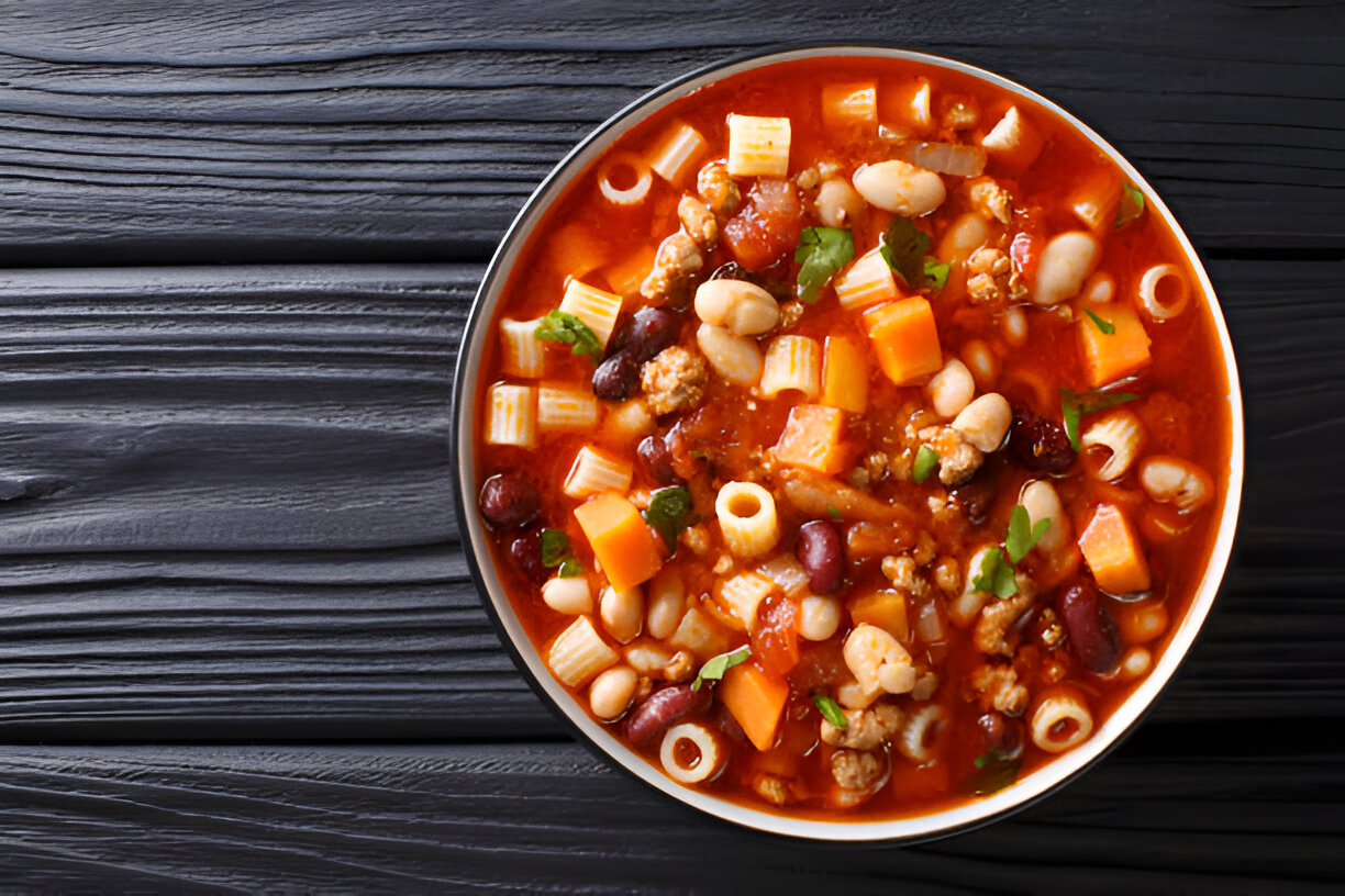 A bowl of colorful mixed bean and pasta soup on a wooden surface.