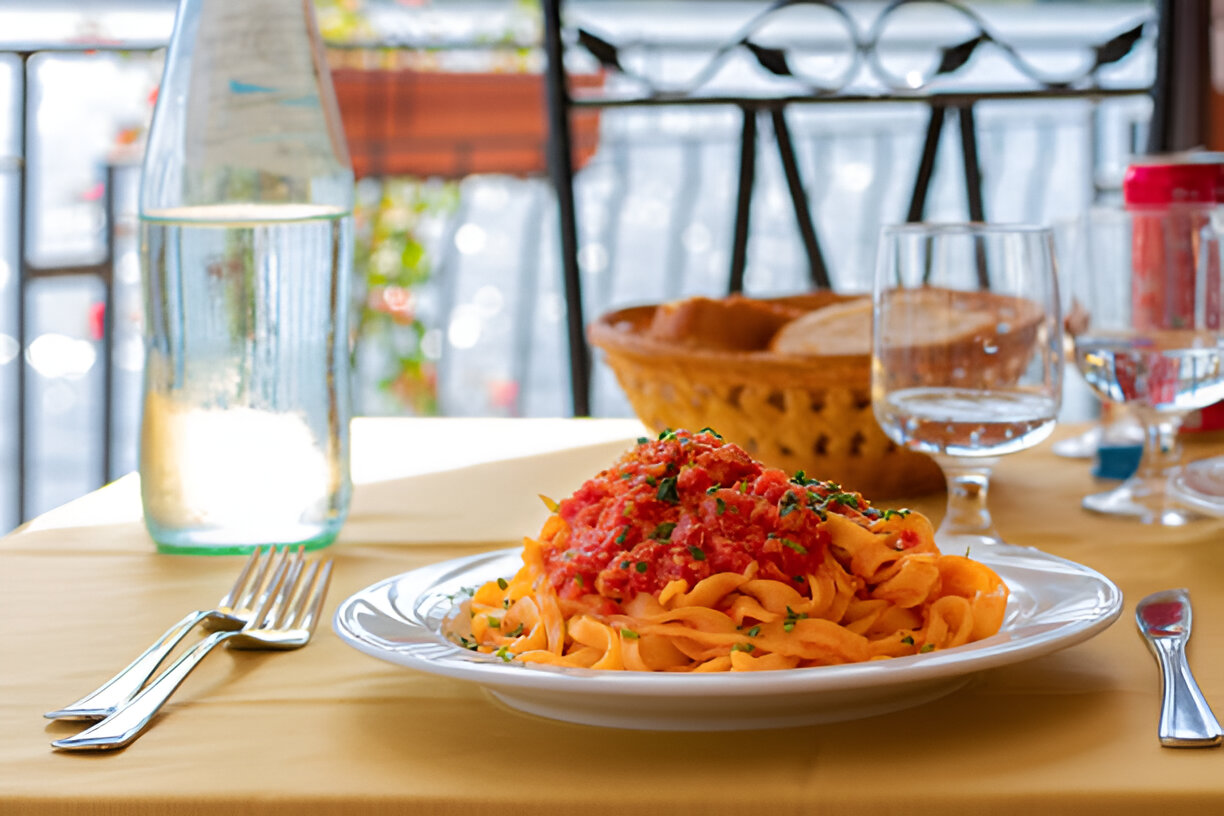 A plate of fettuccine pasta with tomato sauce and herbs on a table with a water bottle and bread basket in a restaurant setting.