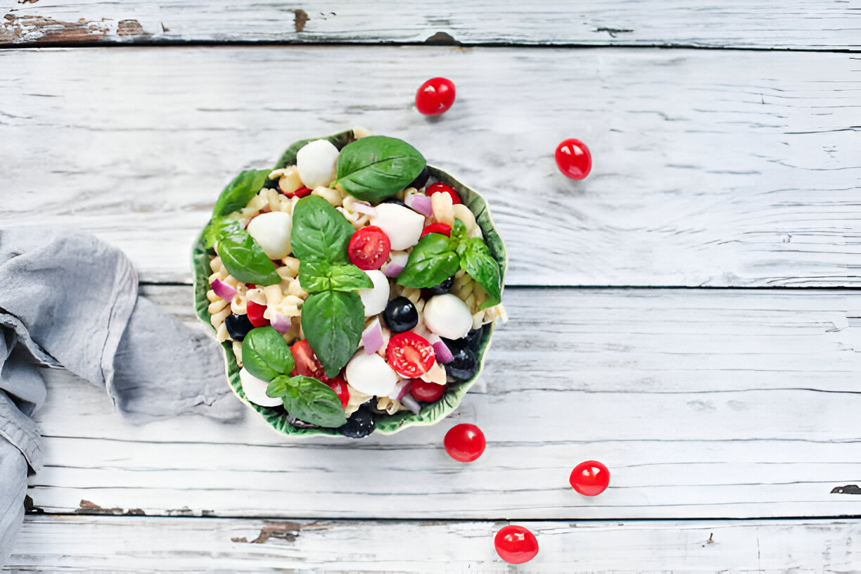 A fresh pasta salad with mozzarella balls, cherry tomatoes, black olives, and fresh basil on a wooden table.