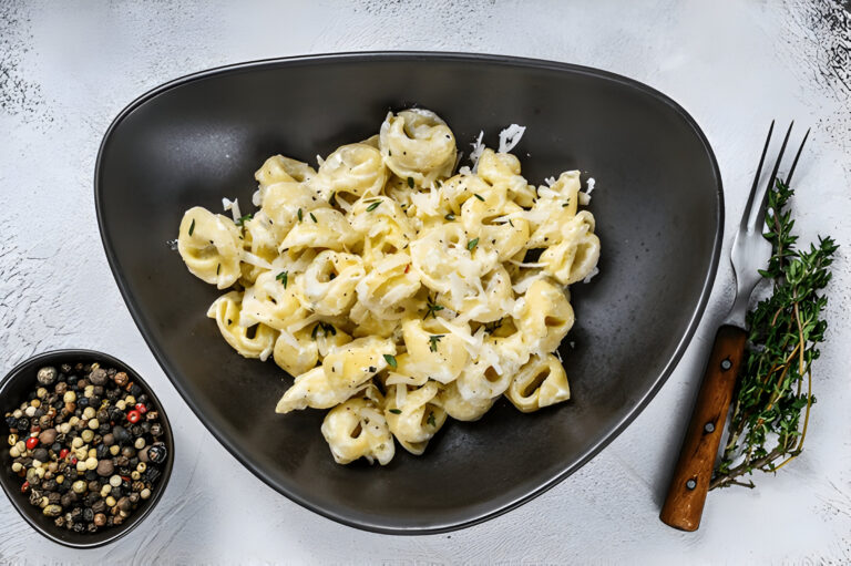 A plate of creamy tortellini pasta with herbs on a textured surface, alongside a bowl of assorted peppercorns and a fork with a wooden handle.