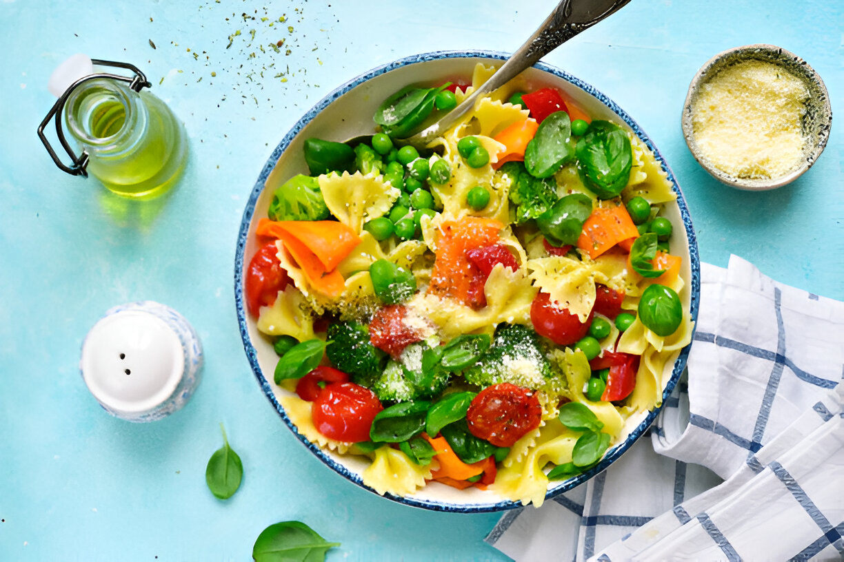 A vibrant bowl of pasta salad with bowtie pasta, cherry tomatoes, green peas, and fresh vegetables, topped with grated cheese and basil.