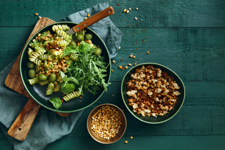 A colorful vegetarian dish featuring spiral pasta, greens, green olives, and a side of popped grains on a wooden table.