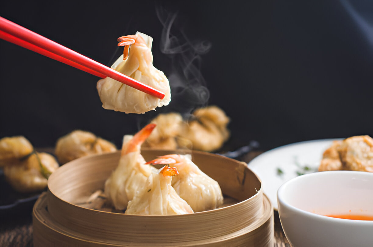 A person holding a steamed dumpling with shrimp using red chopsticks, with steam rising, and a white bowl of dipping sauce in the foreground.