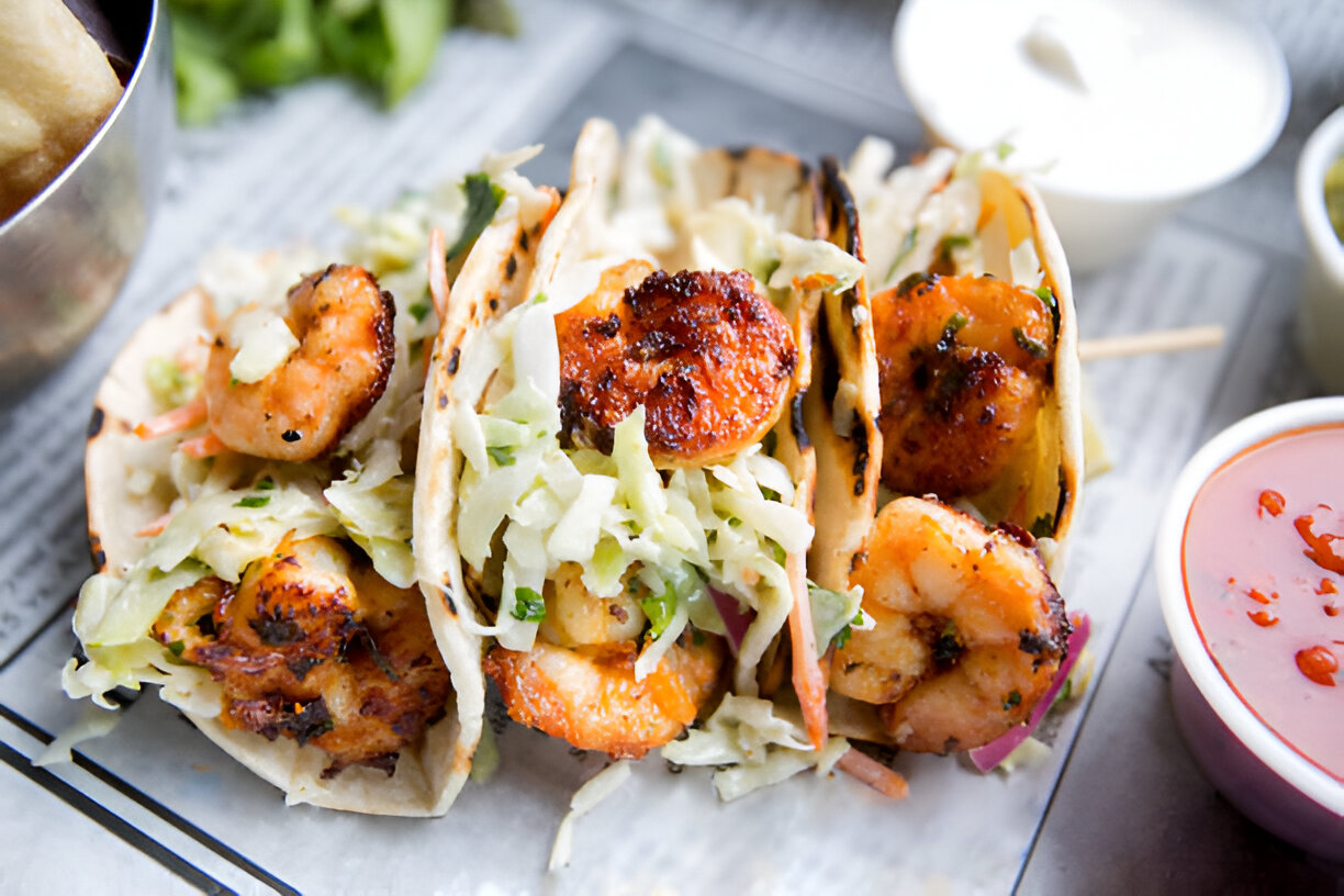 Three shrimp tacos filled with shredded lettuce and served with sauces on the side.