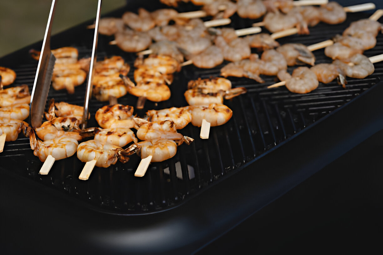 Grilled shrimp skewers on a barbecue grill with tongs lifting one skewer.
