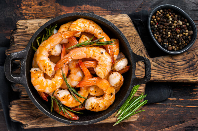 A skillet filled with cooked shrimp garnished with herbs, next to a bowl of mixed peppercorns.