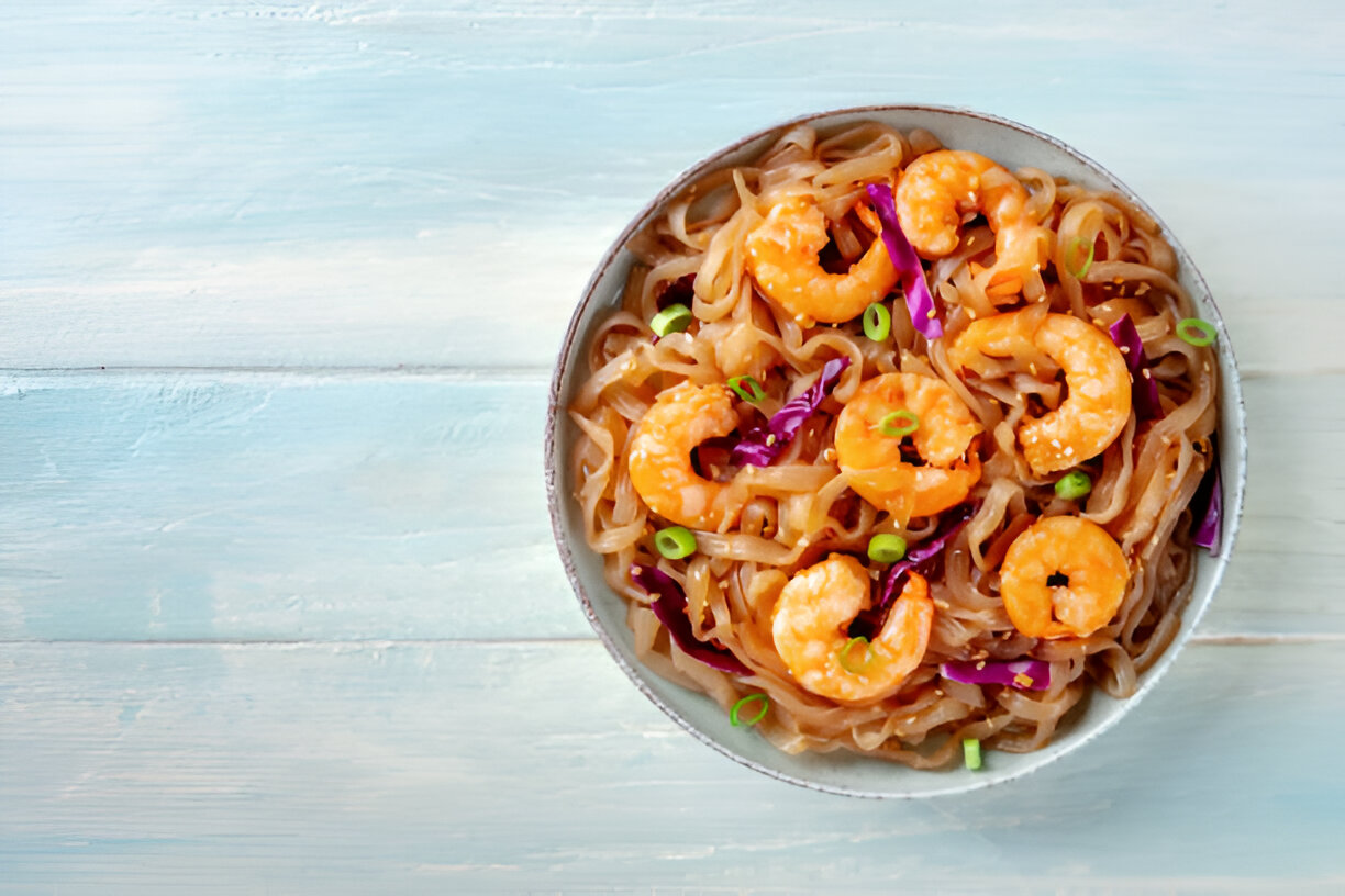 A bowl of shrimp noodles with purple cabbage and green onions on a light blue wooden surface