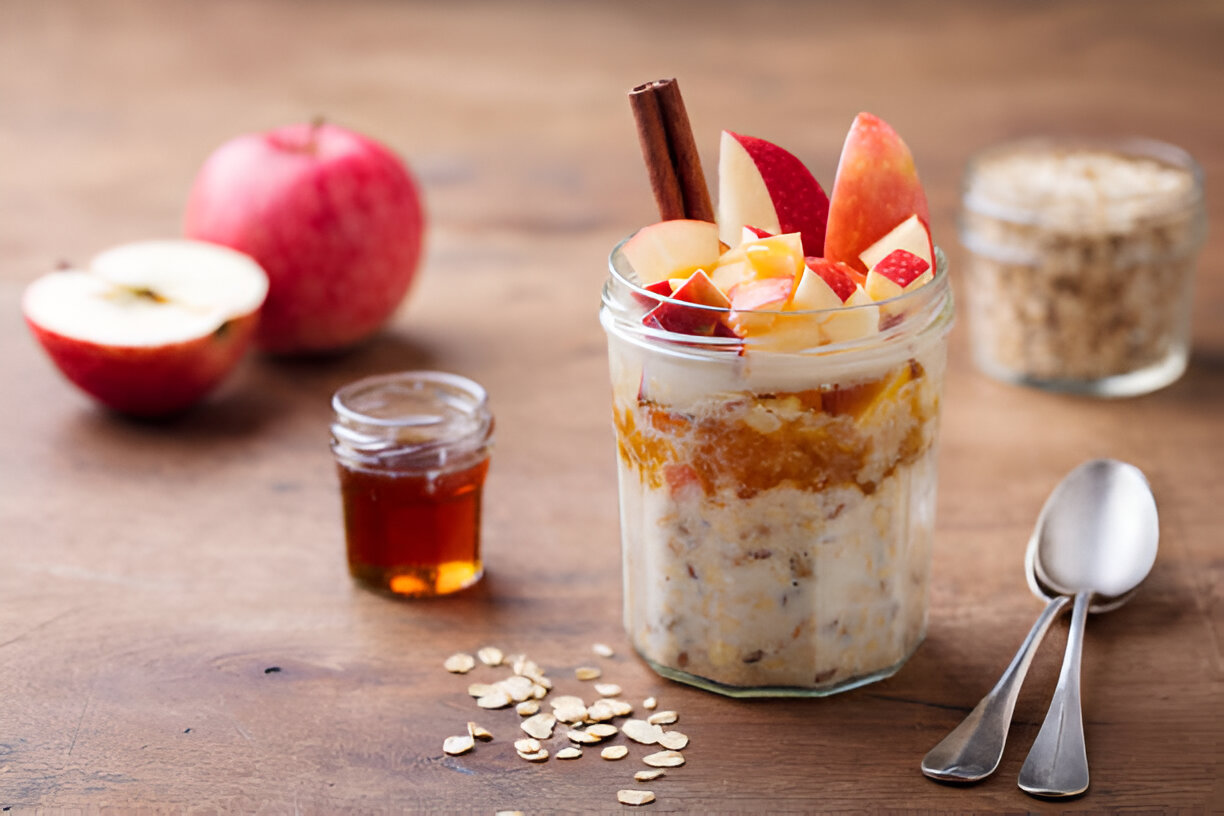 A jar of layered oatmeal with chopped apples, honey, a cinnamon stick, and oats scattered on a wooden table.